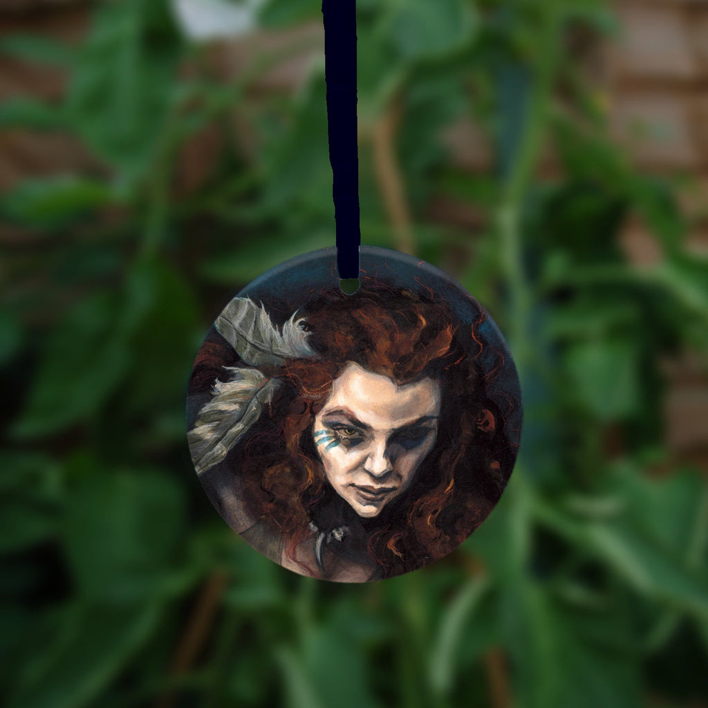 Circular hanging ornament with dark blue ribbon: The head and chest of a woman with long red hair decorated with two feathers, and with lines of blue paint extending away from her eyes.
