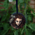 Circular hanging ornament with dark blue ribbon: The head and chest of a woman with long red hair decorated with two feathers, and with lines of blue paint extending away from her eyes.