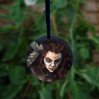 Circular hanging ornament with dark blue ribbon: The head and chest of a woman with long red hair decorated with two feathers, and with lines of blue paint extending away from her eyes.