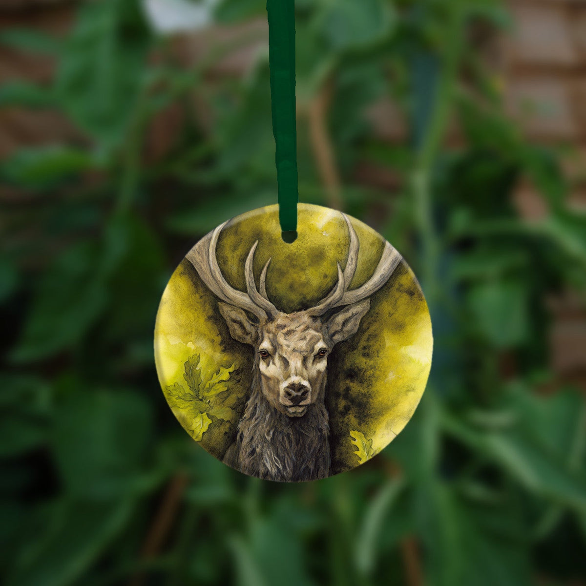 Circular hanging ornament with a green ribbon: The head and chest of an antlered stag directly facing the viewer. Oak leaves are drawn to his left and right. The background is mottled yellows and browns.