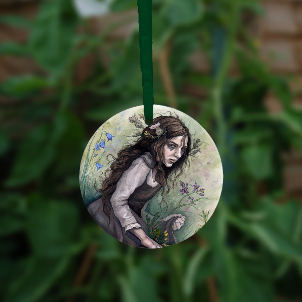 A circular hanging ornament with a green ribbon: A young woman wearing a long brown sleeveless dress with a pale long sleeved top underneath, is surrounded by flowers as she kneels on the ground. The front of her dress is gathered in her hands to form an apron containing sprigs of bog myrtle and yellow gorse flowers. Heather grows next to her. Behind her are harebells, with woody nightshade to the other side and nettles and yarrow to the front. Thistles appear to be tied into her long brown hair.