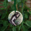 A circular hanging ornament with a green ribbon: A young woman wearing a long brown sleeveless dress with a pale long sleeved top underneath, is surrounded by flowers as she kneels on the ground. The front of her dress is gathered in her hands to form an apron containing sprigs of bog myrtle and yellow gorse flowers. Heather grows next to her. Behind her are harebells, with woody nightshade to the other side and nettles and yarrow to the front. Thistles appear to be tied into her long brown hair.