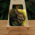 A square coaster: The head and shoulders of a hare facing to the right. Sprigs of red rowan berries and leaves are drawn below. The background is mottled yellows and browns.