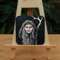 A square coaster: An older woman with blue eyes stands facing the viewer. Her head is covered by a brown hood, and her long grey hair, some of it braided with blue braid, falls down below her face. She holds a staff, topped with a deer antler, upright in one hand. A sea of stars shine behind her.