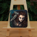 A square coaster: The head and chest of a woman with long red hair decorated with two feathers, and with lines of blue paint extending away from her eyes. The background is teal and green vegetation.