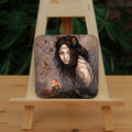 A square coaster: The head and body of a young woman appears above a thick tree branch, upon which sits a red squirrel. The woman is turning her head as if to look behind her. She has long brown hair with leaves stuck in it. Autumnal leaves and branches fill the scene. Colours are reds, oranges and browns.