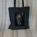 Black tote bag: An older woman with blue eyes stands facing the viewer. Her head is covered by a brown hood, and her long grey hair, some of it braided with blue braid, falls down below her face. She holds a staff, topped with a deer antler, upright in one hand. A sea of stars shine behind her.