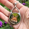A hand holding an oval keyring with a central circular image: The head and neck of a red fox, their face angled towards the front right. The background consists of swirls of branches and leaves in a rich green colour.