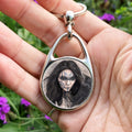 A hand holding an oval keyring with a circular central image: The head and shoulders of a woman with a raven sitting on her shoulder. She has long dark hair and her face is decorated with patterns in blue paint. Her hands are held together against her body. On her wrist she wears a blue band with a raven skull attached. The background is a mottled brown with feathers and swirls drawn within.