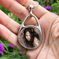 A hand holding an oval metal keyring with a circular central insert: The head and shoulders of a young woman. The woman is turning her head as if to look behind her. She has long brown hair with leaves stuck in it. Autumnal leaves and branches fill the scene. Colours are reds, oranges and browns.