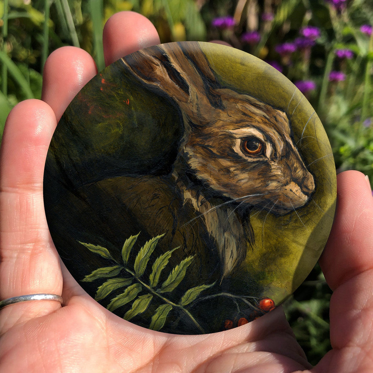 A hand holding a circular object: The head and shoulders of a hare facing to the right. Sprigs of red rowan berries and leaves are drawn below. The background is mottled yellows and browns.