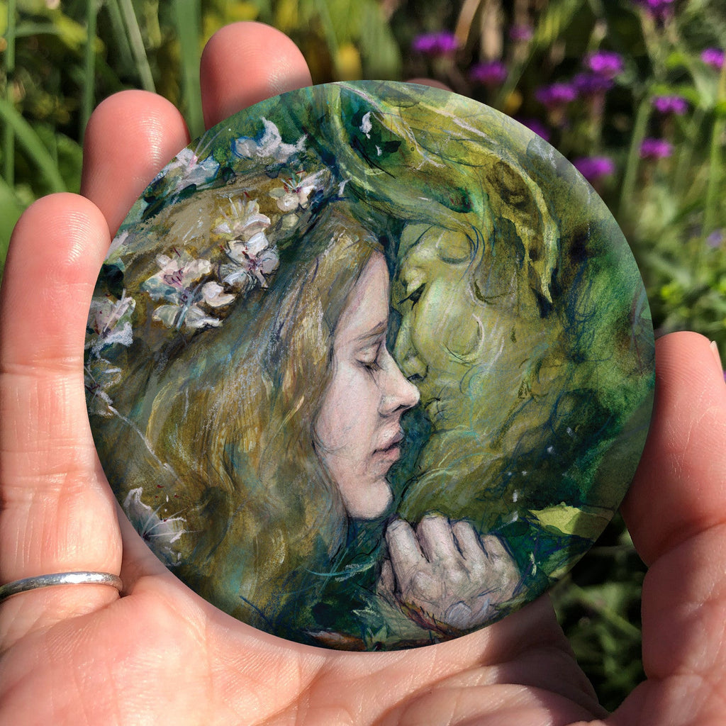 A hand holding a circular object: A young woman with a garland of pale flowers in her hair closes her eyes as she moves to kiss a green man.