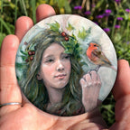 A hand holding a circular object: A young woman looks towards a robin sitting at eye level on the fingers of her hand. Sprigs of red berries and ivy are woven into her hair.