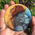 A hand holding a circular object: A painting of two diagonal halves: to the top/left, against a background of mottled orange, a young woman with red hair and orange dress faces to the left. She appears to be leaning against, and blending with, an elderly woman who faces towards the right/bottom in the other half of the design. This half is coloured in shades of blue and grey.