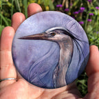 A hand holding a circular object: A sideways view of the head and neck of a heron. The background is a mottled purple with a swirl pattern.