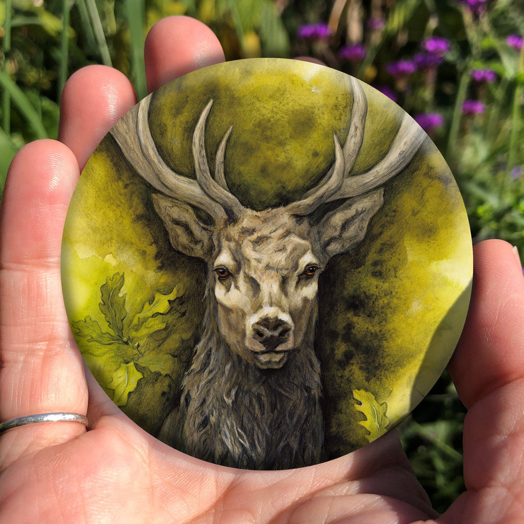 A hand holding a circular object: The head and chest of an antlered stag directly facing the viewer. Oak leaves are drawn to his left and right. The background is mottled yellows and browns.