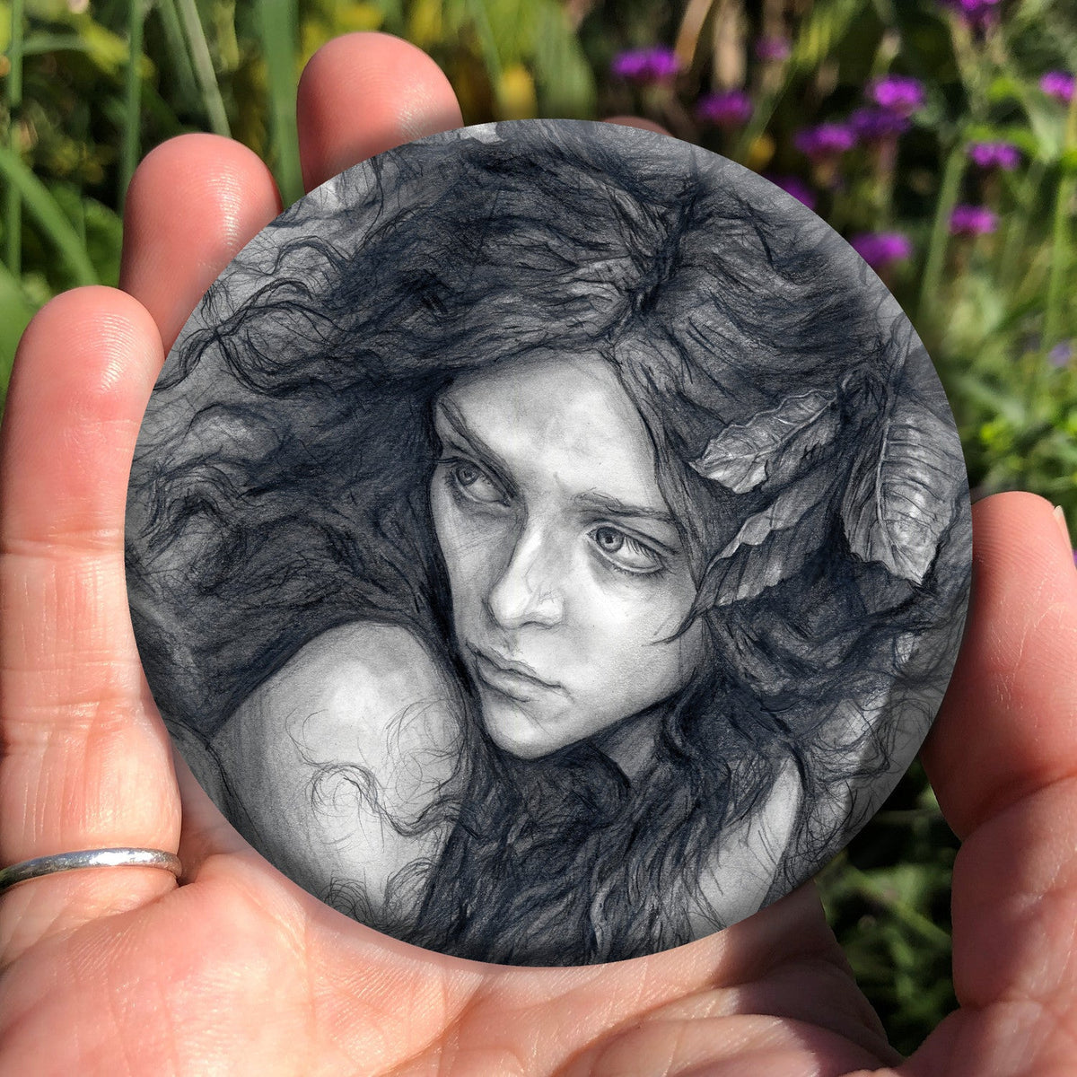 A hand holding a circular object: Pencil illustration of the head and shoulders of a young woman looking back over her shoulder. She has a mass of hair sticking out in all directions around her head and down her front. Leaves are woven - or perhaps stuck - in her hair.