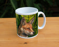 A white ceramic mug: The head and neck of a red fox, their face angled towards the front right. The background consists of swirls of branches and leaves in a rich green colour.