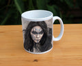 A white ceramic mug: The head and shoulders of a woman with a raven sitting on her shoulder. She has long dark hair and her face is decorated with patterns in blue paint. Her hands are held together against her body. On her wrist she wears a blue band with a raven skull attached. The background is a mottled brown with feathers and swirls drawn within.