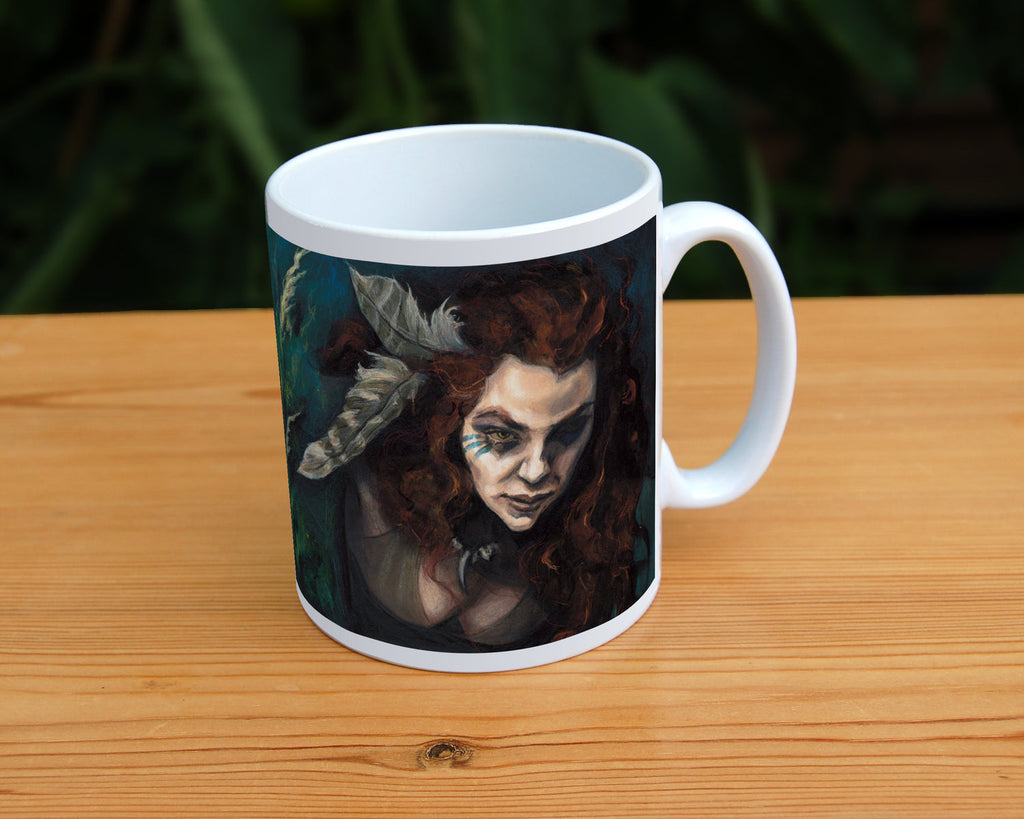 White ceramic mug: The head and chest of a woman with long red hair decorated with two feathers, and with lines of blue paint extending away from her eyes, looms into the picture above the head and body of a snarling brown striped cat. The background is teal and green vegetation. Important: The woman fills one side of the mug, with the cat on the other.