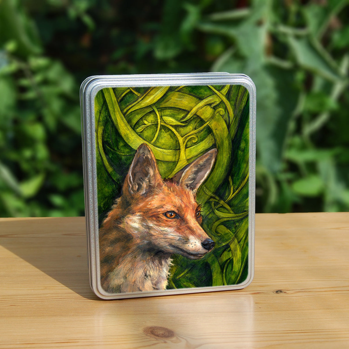 A rectangular tin: The head and neck of a red fox, their face angled towards the front right. The background consists of swirls of branches and leaves in a rich green colour.