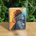 Rectangular metal tin: A painting of two diagonal halves: to the top/left, against a background of mottled orange, a young woman with red hair and orange dress faces to the left. She appears to be leaning against, and blending with, an elderly woman who faces towards the right/bottom in the other half of the design. This half is coloured in shades of blue and grey.