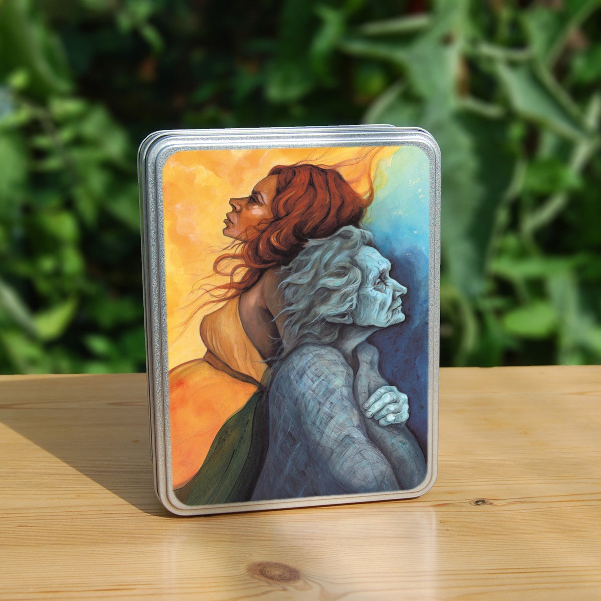 Rectangular metal tin: A painting of two diagonal halves: to the top/left, against a background of mottled orange, a young woman with red hair and orange dress faces to the left. She appears to be leaning against, and blending with, an elderly woman who faces towards the right/bottom in the other half of the design. This half is coloured in shades of blue and grey.