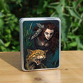 Rectangular tin: The head and chest of a woman with long red hair decorated with two feathers, and with lines of blue paint extending away from her eyes, looms into the picture above the head and body of a snarling brown striped cat. The background is teal and green vegetation.