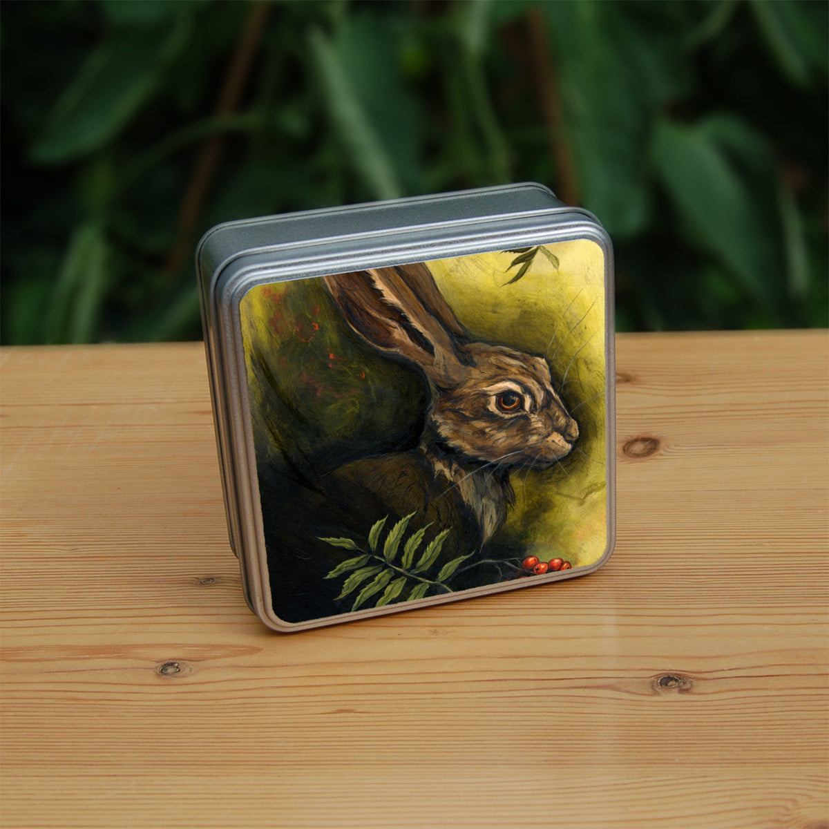 Square metal tin: The head and shoulders of a hare facing to the right. Sprigs of red rowan berries and leaves are drawn below. The background is mottled yellows and browns.