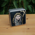 A square tin: An older woman with blue eyes stands facing the viewer. Her head is covered by a brown hood, and her long grey hair, some of it braided with blue braid, falls down below her face. She holds a staff, topped with a deer antler, upright in one hand. A sea of stars shine behind her.