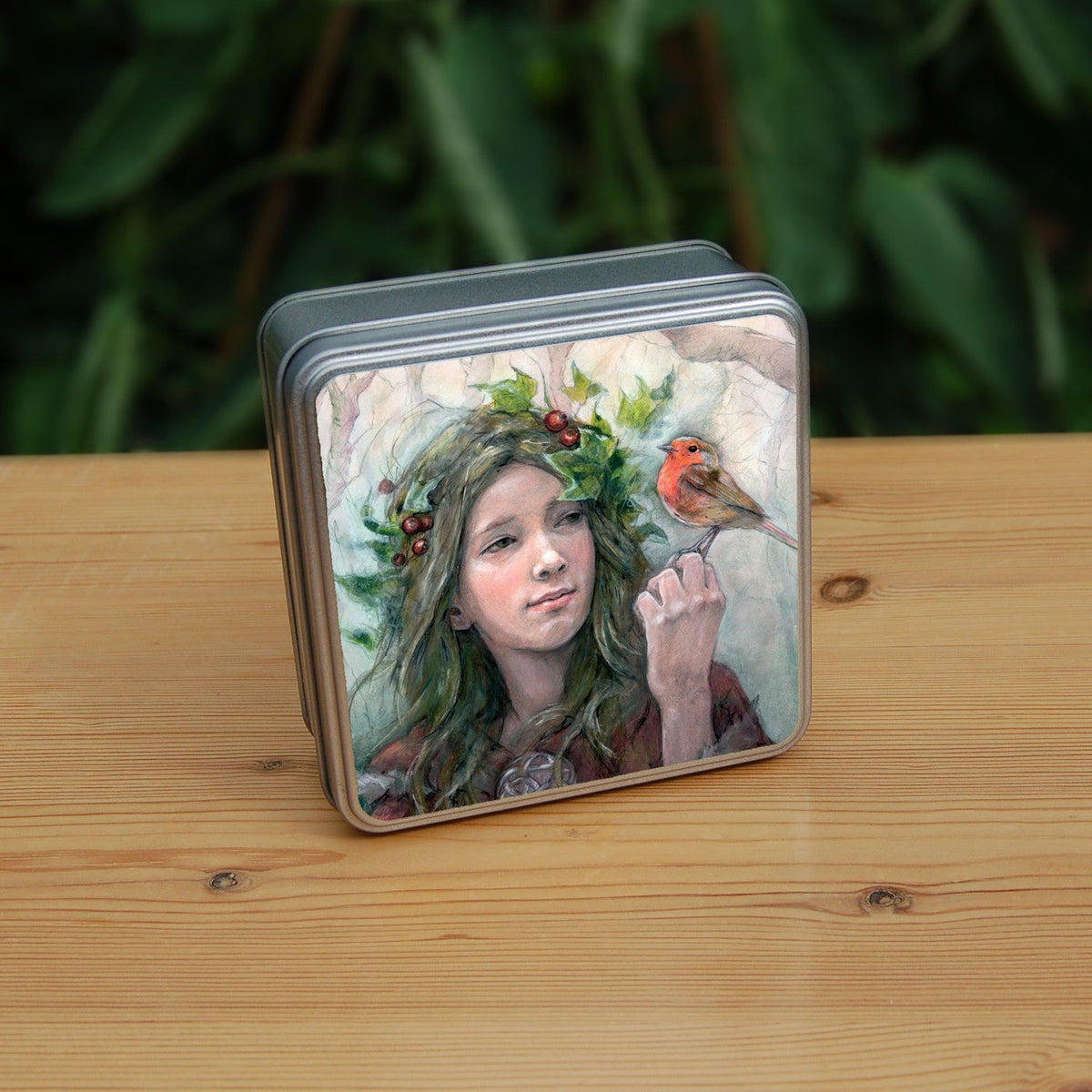 Square metal tin: A young woman looks towards a robin sitting at eye level on the fingers of her hand. She wears a red cloak with white fur trim, closed with a large Celtic brooch. Sprigs of red berries and ivy are woven into her hair. Bare branches of trees fill the background.