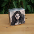 A square tin: The head and shoulders of a woman with a raven sitting on her shoulder. She has long dark hair and her face is decorated with patterns in blue paint. Her hands are held together against her body. On her wrist she wears a blue band with a raven skull attached. The background is a mottled brown with feathers and swirls drawn within.