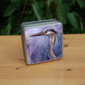 A square tin: A sideways view of the head and neck of a heron. The background is a mottled purple with a swirl pattern.
