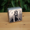 Square tin: A young woman crouches on a pile of stones, her hands resting on stones in front of her. The woman wears a headband with a raven skull attached to the front. A full moon rises behind her, and a silhouette of a hare leaps in front of it. An inscription over the moon reads: Up from the earth with tooth and claw we rise. Stems and flowers fill the rest of the scene.