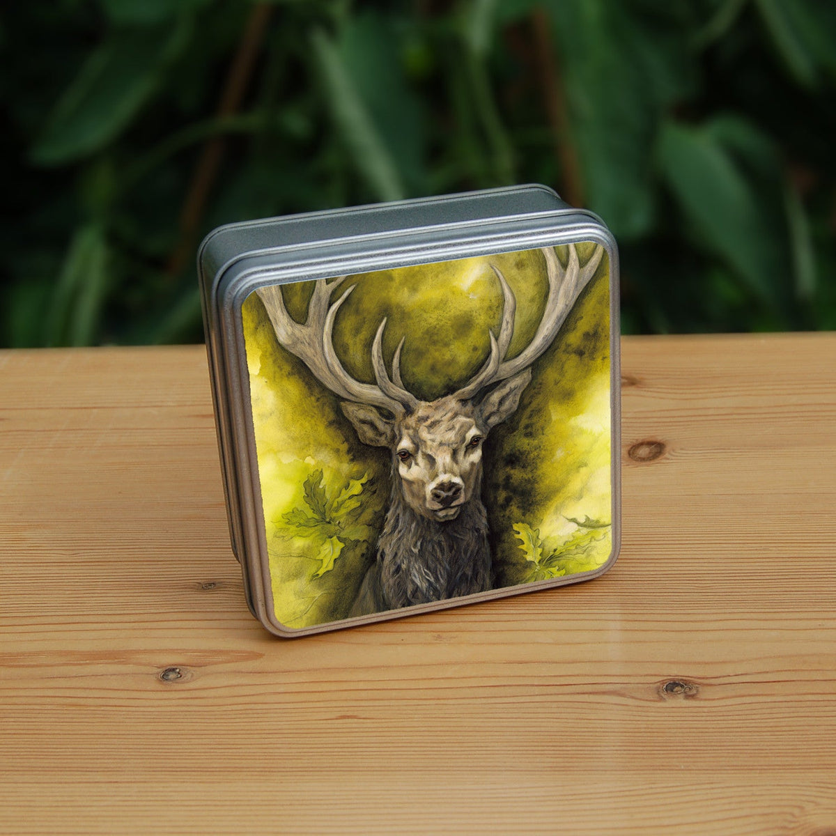 Square metal tin: The head and chest of an antlered stag directly facing the viewer. Oak leaves are drawn to his left and right. The background is mottled yellows and browns.