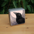 Square tin: The head and body of a raven standing facing towards the left, head tilted upward, behind which is a full moon. The background consists of swirls in a pale pink-brown colour.