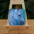 The head and mane of a horse, head facing mostly to the right. The horse is painted entirely in blue apart from a deep brown eye, with two spirals on and above the cheek. The background is blue-green.