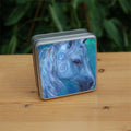 The head and mane of a horse, head facing mostly to the right. The horse is painted entirely in blue apart from a deep brown eye, with two spirals on and above the cheek. The background is blue-green.