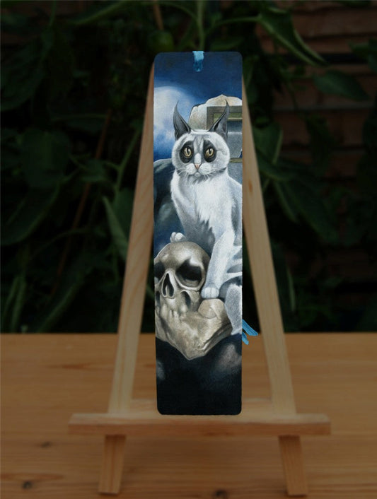 A white cat with black fur around the eyes and black ears sits perched on a skull and looking towards the viewer. In the background is a broken window and a full moon rising from behind clouds. Colours are deep blue, grey and light brown.