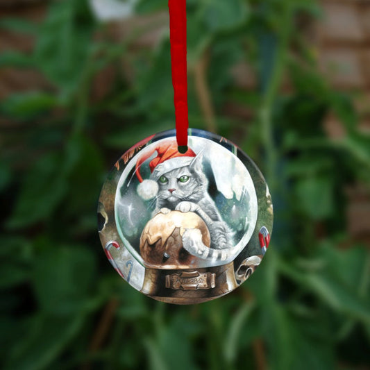 A pale silver tabby cat wearing a santa hat sits in a snow globe, front paws resting on a Christmas pudding. "Krampuss" is written on the globe stand beneath. 