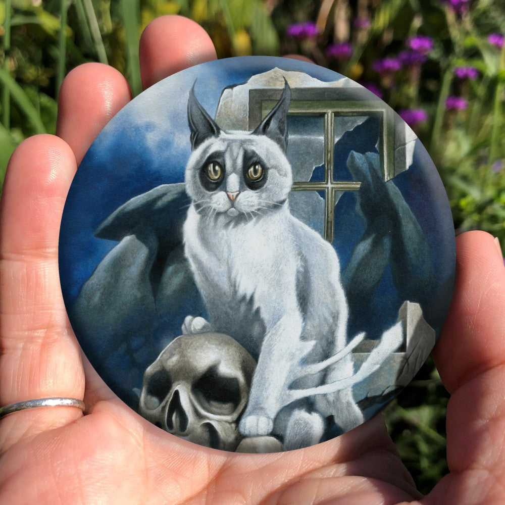 A white cat with black fur around the eyes and black ears sits perched on a skull and looking towards the viewer. In the background is a megalith, a broken window and a full moon rising from behind clouds. Colours are deep blue, grey and light brown.