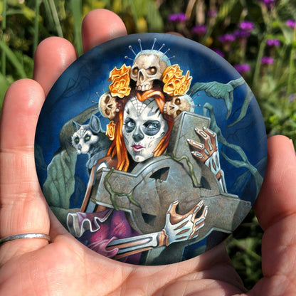 A woman with her face and arms decorated as a skeleton clutches a large stone Celtic cross to her chest. Her red hair is decorated with skulls with pins protruding from their heads. A black cat with a white face sits on her shoulder.