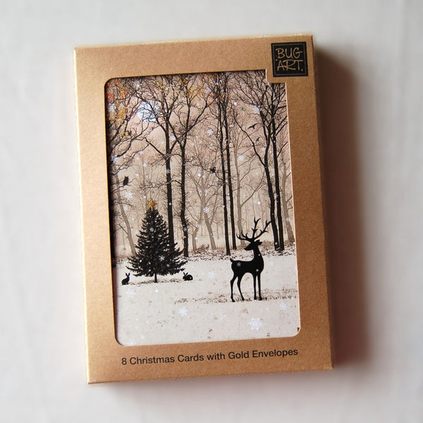 The silhouette of a stag stands in the foreground against a backdrop of bare winter trees. Snow is falling and settling on the ground. A Christmas fir tree stands midway between the trees and stag, a rabbit sits underneath. Colours are mostly browns.