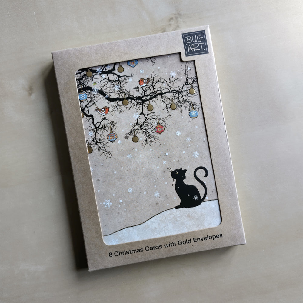 The silhouette of a black cat sitting looking up towards the silhouette of a leafless tree. The tree is decorated with colourful baubles. Two robins sit in the tree, and snow falls all around. The background is shades of beige.