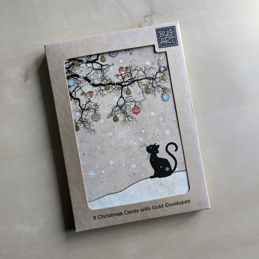 The silhouette of a black cat sitting looking up towards the silhouette of a leafless tree. The tree is decorated with colourful baubles. Two robins sit in the tree, and snow falls all around. The background is shades of beige.