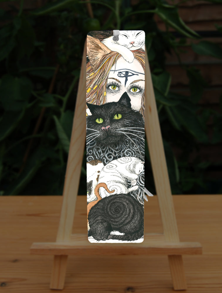 A mass of cats of all colours and in many poses surrounds the face of a red-haired woman with cat-slit eyes and ears, and a Eye of Horus symbol on her forehead. A white cat sits on her head. Colours are oranges, reds, black and white.