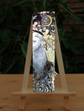 A large grey and white heron stands on one leg in water near the bank of a river. A damselfly hovers nearby. Runes (Berkana and Algiz) dangle from the heron's beak. The river extends behind the scene, with grassland and trees beyond. An unborn human baby lies curled up within the sun.