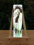 A male figure with long brown hair and beard, both cascading down his body, stands sideways wearing a pale green flowing robe. The side of his head is shaved, and tattooed with a line of characters from the Ogham alphabet. His bound hair contains fungi, a group of feathers, and a mouse. He looks down towards butterflies sitting on his hand. Runes dangle from his arm. A mouse sits in the crook at his elbow. A white stoat appears to form from his shoulder and run down his arm, a badger from his back ...