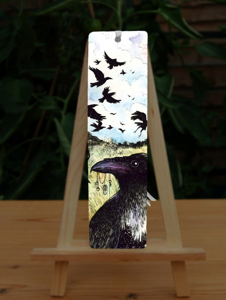 A large flock of ravens sit on and fly around a drystone wall surrounded by yellow-green grassy fields. The blue sky is broken with white clouds. In the foreground a raven holds a string of tokens in its beak, including a pentagram, key and Teiwaz rune.