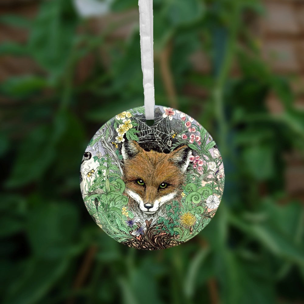 The head of a red fox looks towards the viewer. He's surrounded by a mass of leaves and flowers which rise out from a twisted tree trunk and tangled branches.